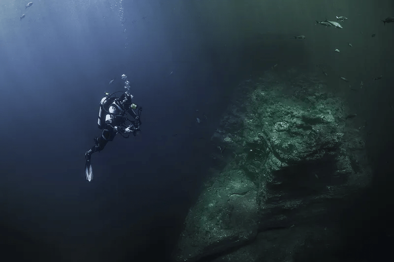 A SCUBA diver is in the center left of the image. The water gets darker at the bottom of the image. In the right of the image is some undersea ruins with a few silver fish