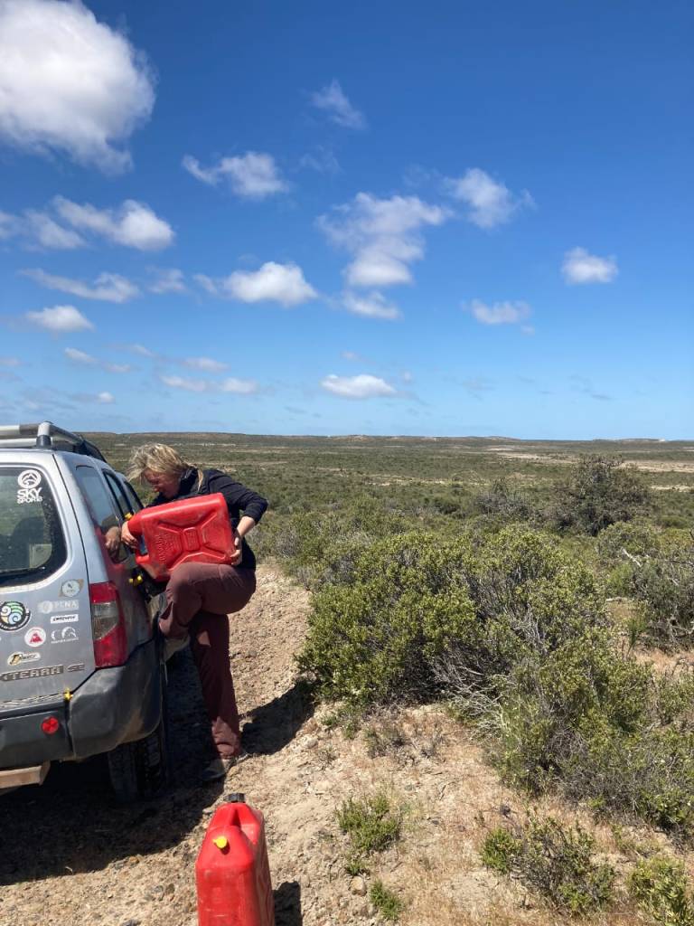 An image of a researcher pouring diesel from large red fuel containers into the gas tank of a truck. The truck is parked on a dry dirt road, surrounded by small scrubby plants stretching for miles in a wide flat landscape, under a blue sky dotted with clouds.
