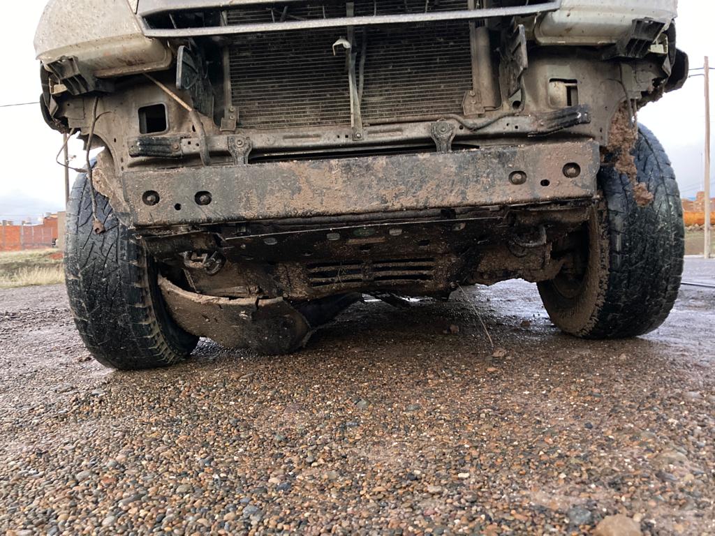 A close-up image of a rugged, extremely muddy truck that the bumper has fallen off of, parked on a gravel road.