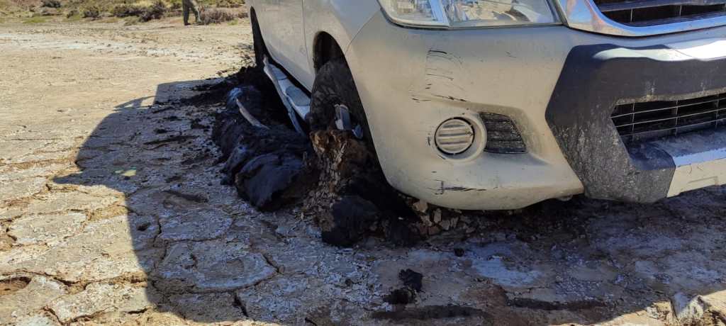 A close-up image of a truck partially sunken into the muddy ground. There are big piles of mud piled up around the wheels of the truck. The surface of the mud is cracked in some places and the mud underneath looks slick and wet.